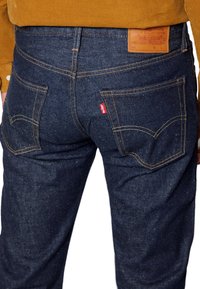 Mörkblå denimjeans med en texturerad yta, med en beige bakficka, gula sömmar och klassisk fickdesign. Röd etikett på vänster ficka.