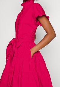 Kvinna klädd i en ljusrosa klänning med korta ärmar, volangkrage, knappdetaljer, midjeband och handen i sidofickan.