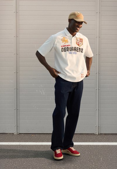 Tricou polo alb cu grafice brodate, blugi albastru închis și adidași roșii cu tălpi crem, asortați cu o șapcă bej.