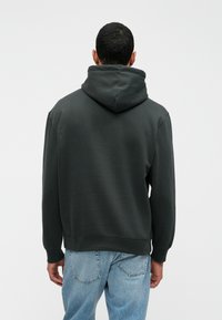 Felpa grigia scura con cappuccio ampio, polsini e orlo a coste. Il tessuto ha un aspetto morbido e strutturato, abbinato a jeans azzurri chiari.