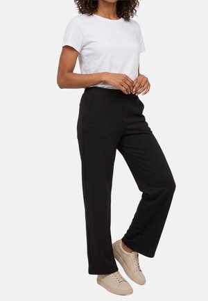 Una persona con una camisa blanca de manga corta, pantalones negros de pierna recta y zapatillas beige, de pie sobre una pierna.