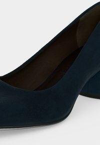 Gros plan d’une chaussure à talon haut en suède bleu foncé montrant un bout arrondi et une doublure intérieure sur un fond clair.