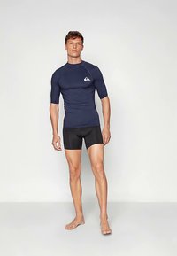 Tengerészkék rövid ujjú vízálló felső kis fehér logóval, fekete fürdőnadrággal párosítva. Az anyag sima és rugalmas hatású.