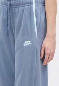 Κοντινός πλάνος ενός ατόμου που φοράει ανοιχτό μπλε φλις παντελόνι Nike με κορδόνι στη μέση και πλαϊνές τσέπες, δείχνοντας το δεξί του χέρι χαλαρό στο πλάι.