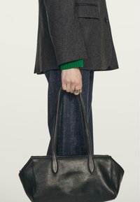 Czarna skórzana torebka o gładkiej teksturze, wydłużonych uchwytach i trójkątnym kształcie, trzymana przez osobę ubraną w szary żakiet i zielony sweter.