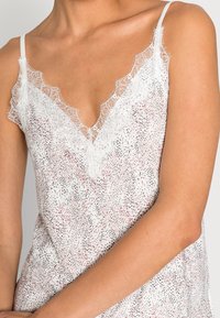 Žena oblečená v bílém topu s krajkovým lemem, pokrytém růžovým a černým puntíkovaným vzorem, zachycena od ramen po pas na jednobarevném pozadí.