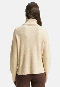 Kobieta w kremowym, prążkowanym swetrze z golfem i brązowych spodniach, stoi tyłem na jednolitym tle.