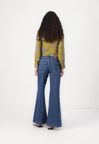 Blusa verde de manga longa com padrões de estrelas, combinada com jeans de ganga azul de perna larga. A vista traseira mostra detalhes dos bolsos e costuras.