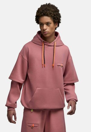 Jeune personne aux cheveux bouclés portant un sweat à capuche ample rose poudré avec des cordons orange et un pantalon assorti avec des détails de poche.