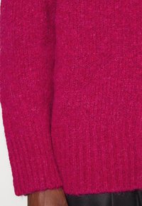 Světlý růžový pletený svetr s texturovaným povrchem, žebrovanými manžetami a volným střihem. Tkanina vypadá měkce a teple s mírně chlupatým povrchem.