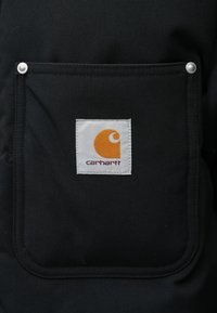 Un bolsillo de tela negra que presenta una etiqueta blanca rectangular con un logo naranja y el texto "carhartt" debajo. Dos broches plateados en el borde.