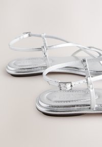 Sandale plate argintii, cu bretele subțiri și finisaj metalic neted. Talpa este plată și ușor căptușită, cu un design minimalist.