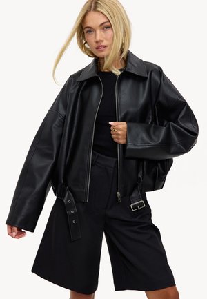 Veste courte en cuir synthétique noir avec silhouette surdimensionnée, grand col et fermeture éclair. Comprend un détail de ceinture latérale et une coupe ample.