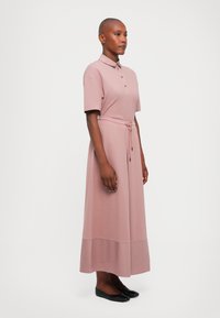 Polo-stil kjole i lys rosa, korte ermer, knapplacket, livrem, og ankellang hem. Glatt stoff og avslappet passform. Svarte leiligheter brukt.