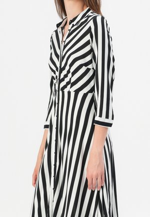 Femme portant une robe à rayures verticales noir et blanc, boutonnée, avec manches trois-quarts sur fond blanc uni.