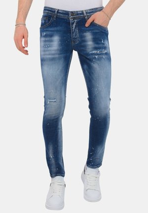Man draagt slim fit versleten blauwe jeans en witte sneakers, met één hand in de zak en zichtbare witte top.