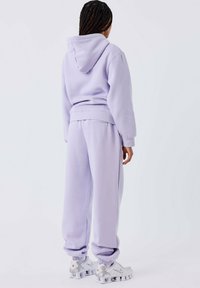 Lila hoodie en bijpassende sweatpants, beide gemaakt van zacht materiaal. Voorzien van een trekkoord in de capuchon, geribbelde manchetten en een relaxte pasvorm. Kenmerkende sneakers te zien.