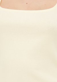 Κοντινή λήψη ενός ατόμου που φοράει ριμπ πλεκτό, αμάνικο, εκρού τοπ με τετράγωνο ντεκολτέ, που δείχνει το πάνω μέρος του στήθους και τους ώμους.