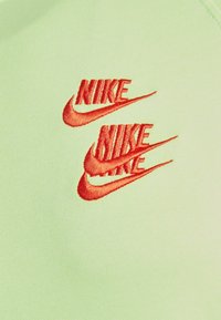 Tři překrývající se červené loga Nike vyšitá na světle zelené látce.