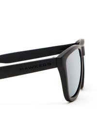 Schwarze Sonnenbrille mit mattem Finish, rechteckigen Gläsern und verspiegelt grauem Farbton. Mit dem Logo "HAWKERS" an der Seite.