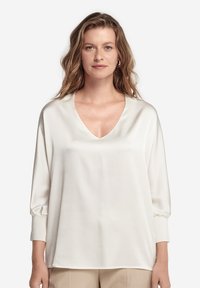 Blusa bianca in seta con scollo a V profondo, spalle scese e maniche lunghe. Il tessuto ha una consistenza liscia e una vestibilità comoda.