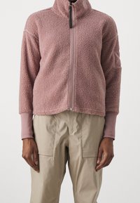 Chaqueta de forro polar rosa claro con cremallera y cuello alto, que presenta mangas largas y una superficie texturizada y suave. Combinada con pantalones cargo beige.