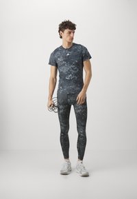 Svart og grå abstrakt mønster på sporty skjorte og leggings laget av elastisk materiale, kombinert med hvite joggesko og holder hodetelefoner.
