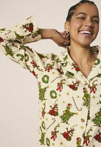 Camisa de pijama color crema con un divertido patrón navideño que incluye elementos en verde y rojo, como árboles, renos y bastones de caramelo.