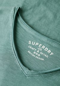Teal v-hals t-shirt lavet af blødt stof, med et minimalistisk logo i hvid, og synlig syning langs halsudskæringen.