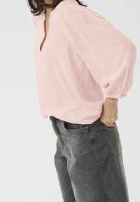 Pink tekstureret bluse med V-udskæring og puffede ærmer, parret med mørkegrå højtaljede jeans med lommer.