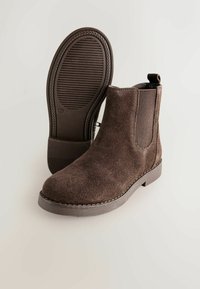 Las botas de tobillo de ante marrón cuentan con paneles laterales elásticos, un tirador y una suela de goma texturizada con surcos para mayor agarre.