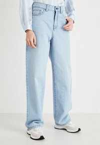 Jeans de pernas largas em azul claro, feito em denim com cintura alta. Apresenta um design clássico de cinco bolsos e fecho de botão. Combinado com ténis brancos.