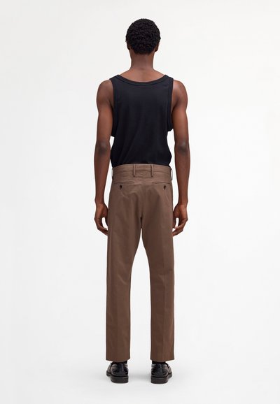 Homme de dos portant un débardeur noir, un pantalon marron et des chaussures noires, sur un fond blanc uni.