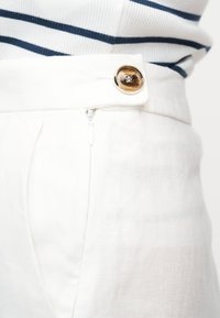 Κοντινό πλάνο λευκών παντελονιών ψηλόμεσων με καφέ κουμπί και φερμουάρ, φορεμένα με λευκό και ναυτικό ριγέ τοπ.