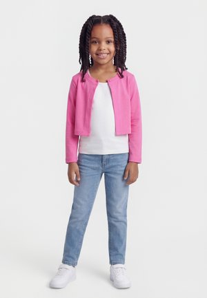 Jeune fille aux cheveux tressés portant un cardigan rose, une chemise blanche, un jean bleu et des baskets blanches, debout et souriante.