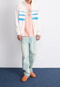 Vit quiltad bomberjacka med blå ränder, rosa grafik T-shirt, urtvättade ljusblå jeans och orange sneakers med gröna detaljer.