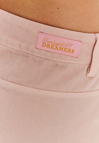 Světle růžová tkanina s vertikálním žebrováním, na které je nálepka s nápisem "Sunlight DREAMERS" zlatým písmem. Jednoduchý a texturovaný design.