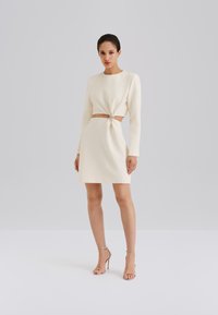 Robe blanche à manches longues avec un détail noué à la taille et un accent argenté. Tissu lisse, longueur genou, coupe ajustée. Associée à des talons argentés.