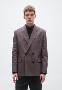 Doppelreihiger Blazer aus einem gedämpften Mauve-Stoff, mit schwarzen Knöpfen, scharfen Revers und zwei Seitentaschen. Über einem schwarzen Hemd getragen.