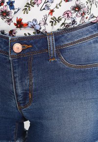 Vaaleansininen farkku, jossa oranssit kontrastisaumat ja metallinappi vyötäröllä. Kukallinen toppi näkyy osittain yläosassa.