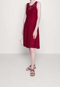 Burgundy spetsklänning med ärmlös design, rund halsringning och utsvängd kjol. Buren med bruna sandaletter med remmar. Ljusrosa handväska som accessoar.