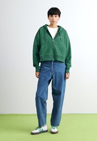 Green zip-up hoodie with front pockets, paired with high-waisted blue denim jeans. Model wears white and green sneakers. Plain background.
