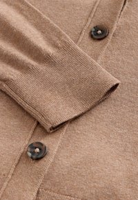 Cardigan en maille marron clair avec une finition texturée, une fermeture à boutons et des poignets côtelés. Le tissu semble doux et léger.