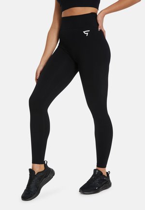 Black high-waisted leggings with a smooth texture, featuring a small white logo on the hip and a fitted silhouette. Worn with black athletic shoes.