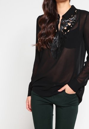 Kvinde iført en sort gennemsigtig bluse med dekorative læderkrave-detaljer og mørkegrønne bukser, stående med den ene hånd i lommen.