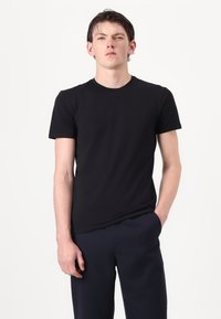 Juodas medvilninis marškinėliai su apvaliu kaklu, trumpomis rankovėmis ir prigludusia struktūra. Dėvimi su tamsiai mėlynomis kelnėmis, fone balta.