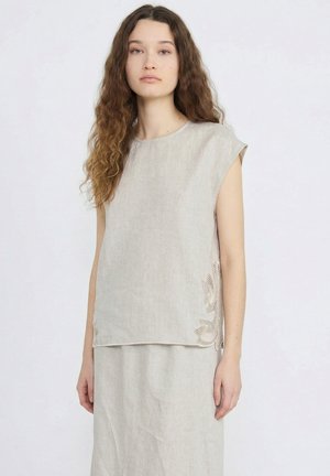 Jeune femme aux longs cheveux bouclés portant un haut beige sans manches et une jupe assortie avec des détails en dentelle sur les côtés.
