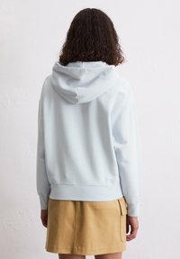 Lichtblauwe hoodie met een relaxed fit, zachte textuur, ribgebreide mouwen en onderrand. Draagt over een tan kleurige rok met zakken.