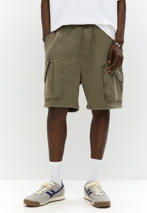 Homme portant un short cargo vert olive avec cordon de serrage, chaussettes blanches, baskets grises et chemise blanche, debout devant un fond blanc.