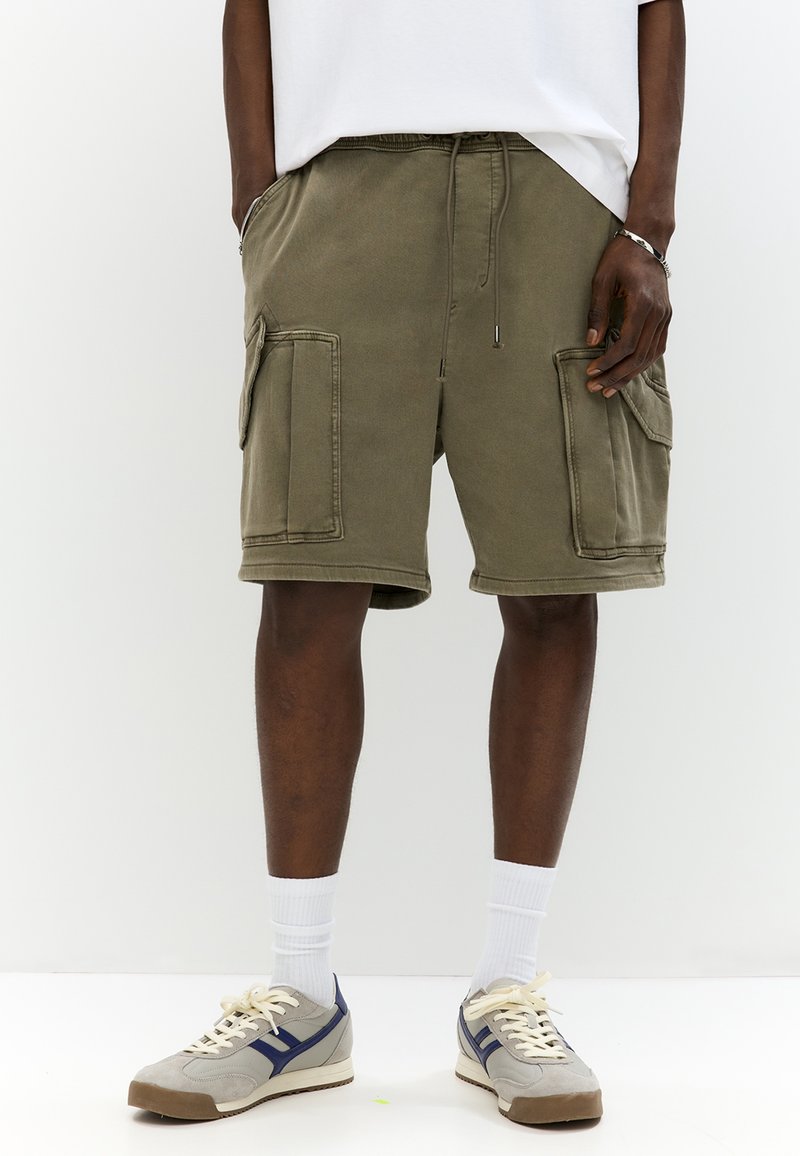 Homme portant un short cargo vert olive avec cordon de serrage, chaussettes blanches, baskets grises et chemise blanche, debout devant un fond blanc.